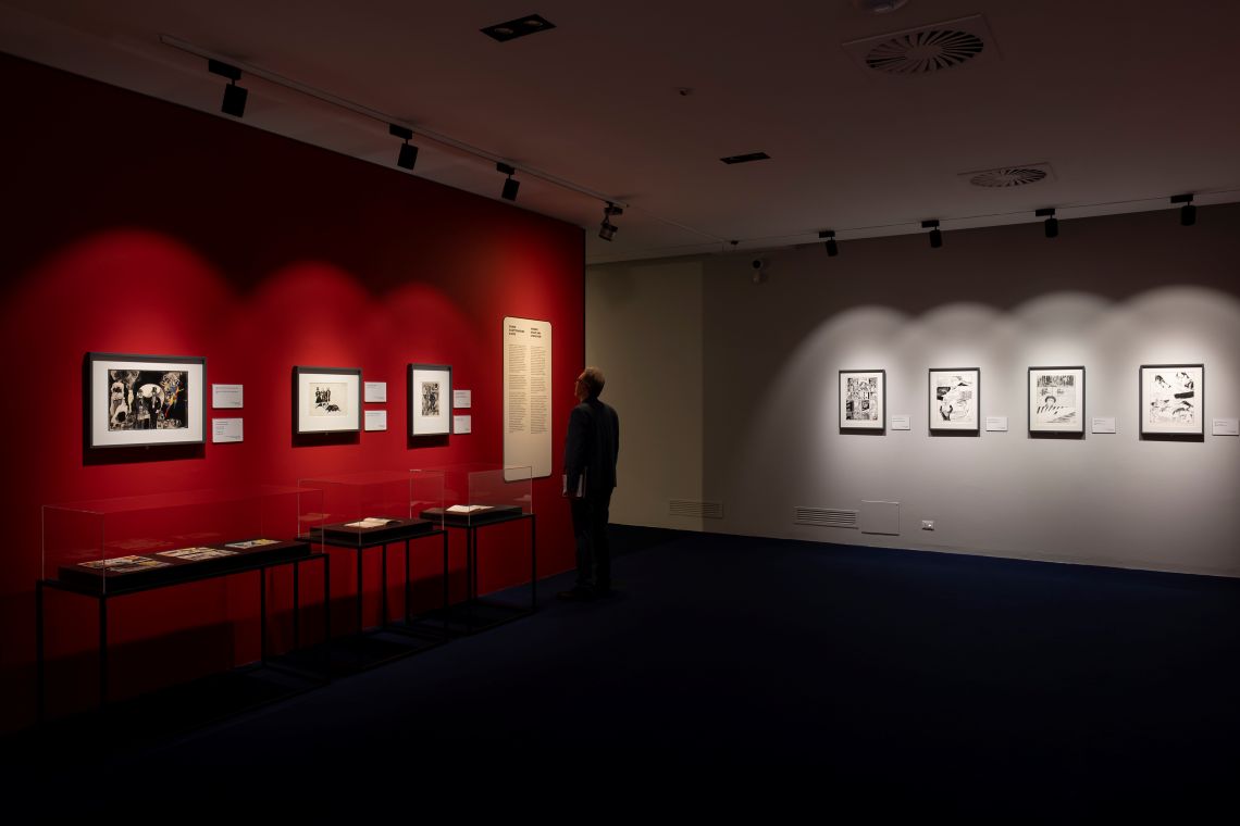 sala con pareti di colore rosso e di colore grigio cui sono appesi diversi disegni e tavole e lateralmente alcune vetrine custodiscono altre opere